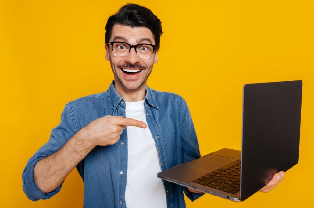 Tráfego pago - Jovem empresario sorrindo e apontando com a mao direita o notebook que está segurando com a mão esquerda