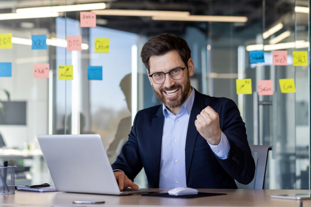 Como criar um site profissional para contadores de sucesso Empresário animado comemorando o sucesso no notebook em um escritório moderno com notas de post-it na parede de vidro